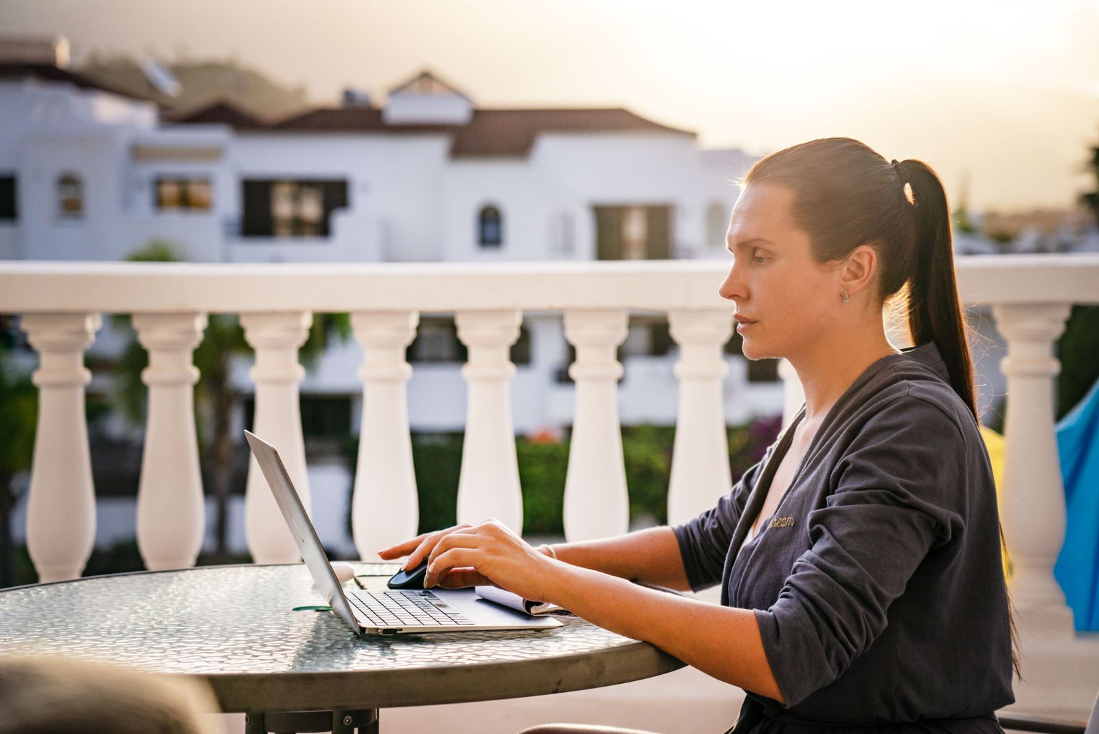 A woman working on a laptop at a table on a sunny villa balcony